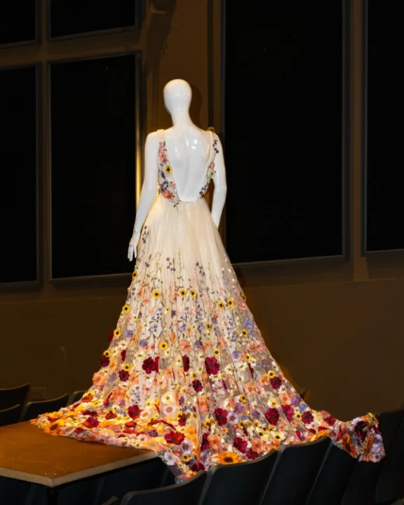 Mannequin facing away from viewer wearing a long, white gown embroidered with an explosion of colorful flowers.