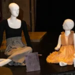 Mother and daughter seated mannequins. Mother wears a black and grey dress. Child wears a pink dress.